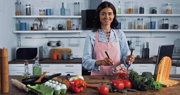Mulher alegre misturando salada de legumes frescos e sorrindo para a câmera na cozinha — Fotografia de Stock