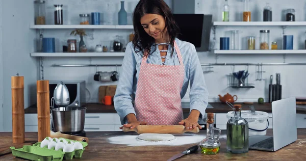 Heureuse femme au foyer dans tablier déroulant la pâte près du robot culinaire et ordinateur portable sur le plan de travail — Photo de stock