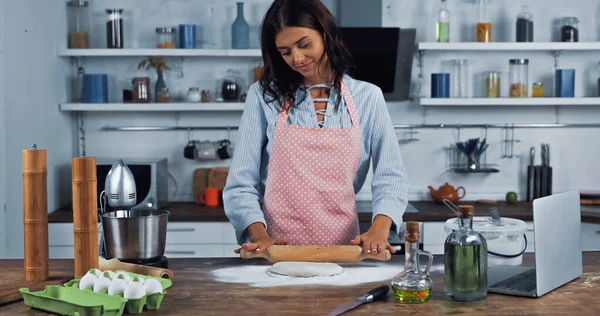 Happy housewife in apron rolling out dough near food processor and laptop on worktop - foto de stock