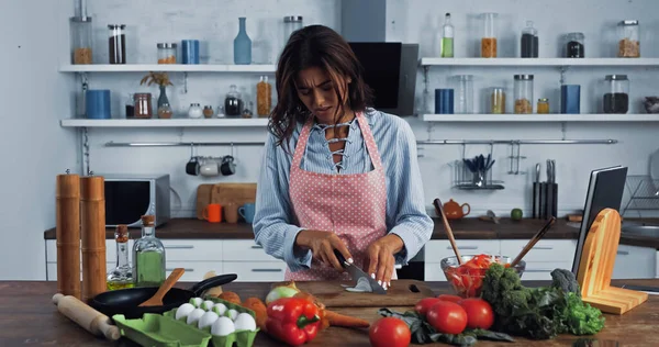 Morena ama de casa cortar cebolla cruda cerca de varias verduras y huevos en la mesa de la cocina - foto de stock