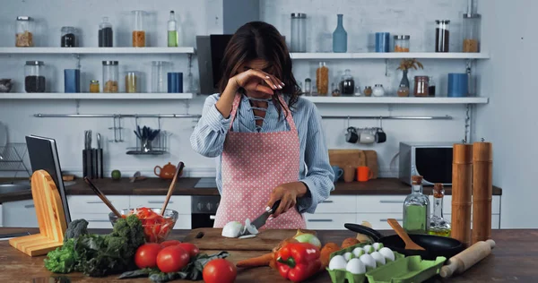 Woman crying and wiping irritated eyes while cutting onion near fresh vegetables — Photo de stock
