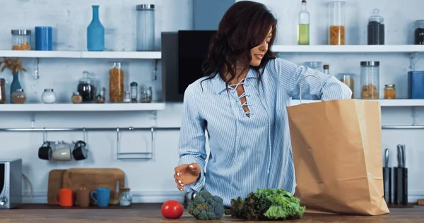 Sorrindo mulher morena perto de saco de papel e legumes frescos na bancada da cozinha — Fotografia de Stock