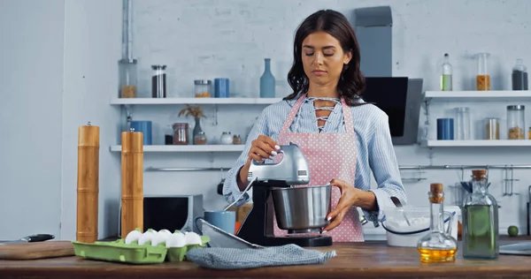 Morena ama de casa utilizando mezclador eléctrico mientras cocina en la cocina - foto de stock