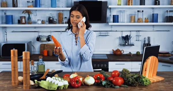 Ama de casa sosteniendo zanahoria fresca y hablando en el teléfono inteligente cerca de las materias primas y libro de cocina - foto de stock