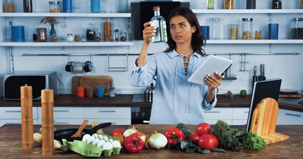 Dona de casa com tablet digital olhando para garrafa com óleo perto de ingredientes crus na cozinha — Fotografia de Stock