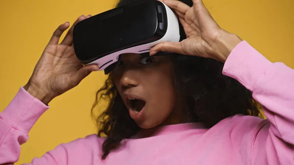 Shocked african american woman in pink sweatshirt holding vr headset isolated on yellow - foto de stock