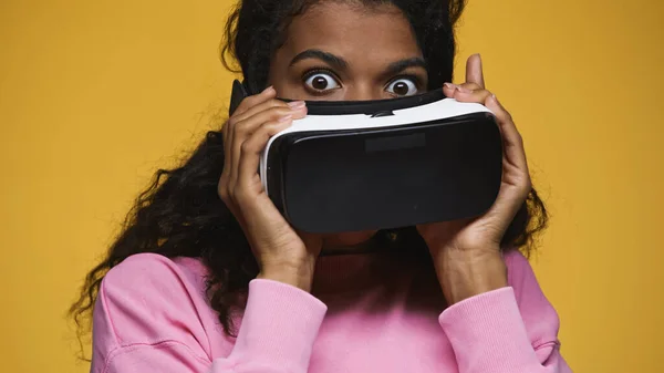 Surprised african american woman in pink sweatshirt holding vr headset isolated on yellow - foto de stock