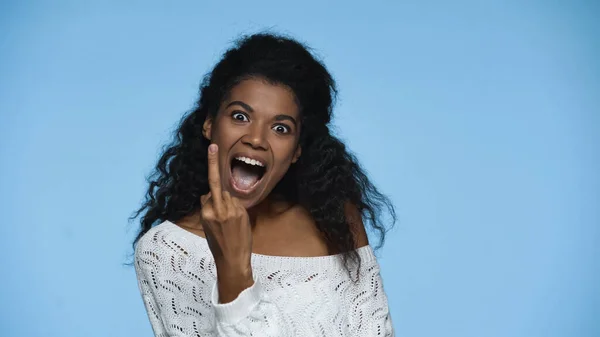 Happy african american woman in white knitted sweater showing middle finger isolated on blue — Stockfoto