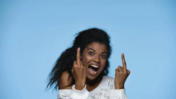Amazed african american woman in white knitted sweater showing middle fingers isolated on blue — Stockfoto