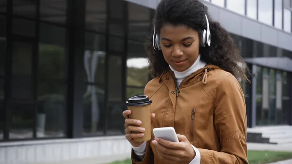 Encaracolado mulher americana africana em fones de ouvido sem fio segurando smartphone e copo de papel fora — Fotografia de Stock