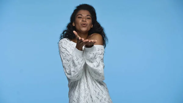 Curly african american woman in white knitted sweater sending air kiss isolated on blue — Stock Photo