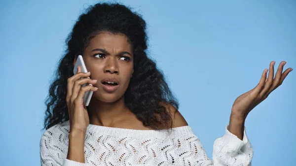 Mulher afro-americana descontente em tricô suéter gestos enquanto fala no celular isolado no azul — Fotografia de Stock