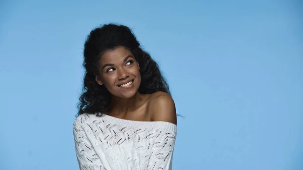 Smiling african american woman in white knitted sweater looking away and biting lip isolated on blue — Stockfoto