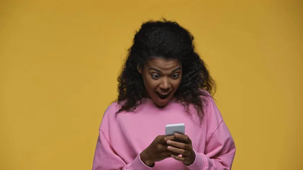 Amazed african american woman in pink sweatshirt chatting on smartphone isolated on yellow — стоковое фото