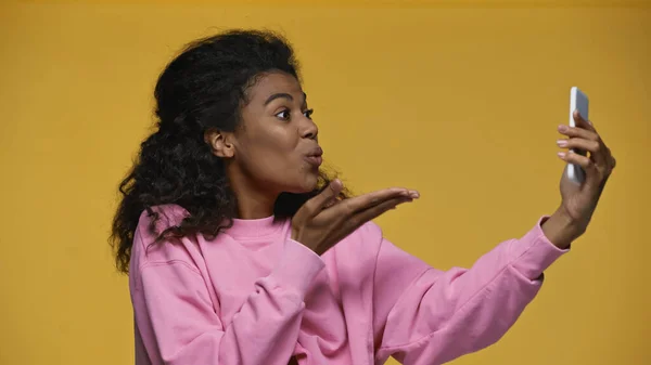 African american woman in pink sweatshirt sending air kiss while taking selfie isolated on yellow — стокове фото