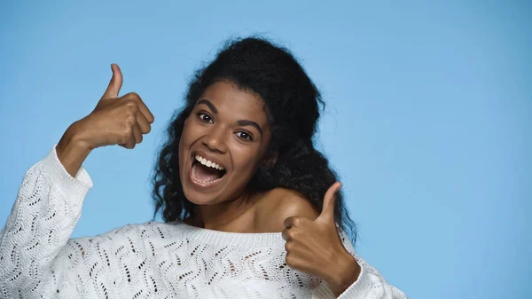 Cheerful african american woman in white knitted sweater showing thumbs up isolated on blue — Stock Photo
