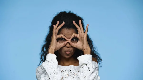 African american woman in knitted sweater grimacing and showing hand gesture symbolizing binoculars isolated on blue — Photo de stock