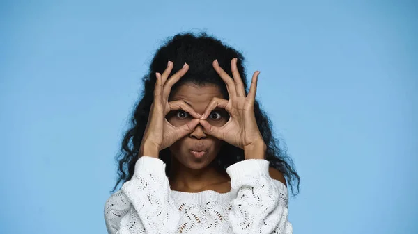 Mulher afro-americana em camisola de malha sorrindo e mostrando gesto de mão simbolizando binóculos isolados em azul — Fotografia de Stock