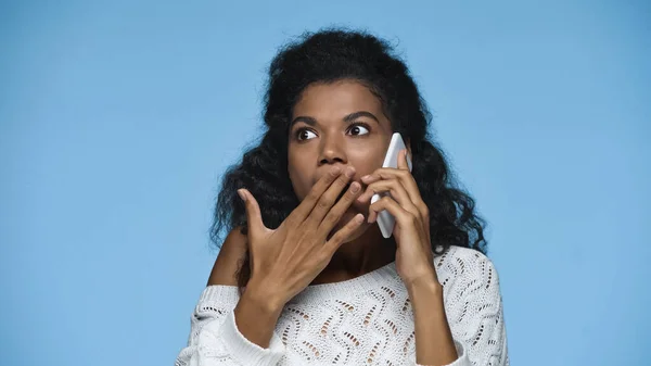 Chocado afro-americano mulher em malha camisola cobrindo boca enquanto fala no smartphone isolado no azul — Fotografia de Stock