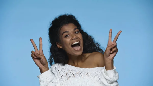 Amazed african american woman in white knitted sweater showing peace sign isolated on blue — Stockfoto
