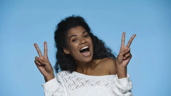 Amazed african american woman in white knitted sweater showing peace sign isolated on blue — Photo de stock