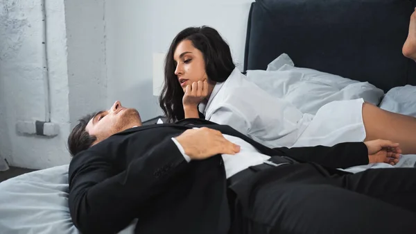 Businessman in suit lying on bed and talking with girlfriend in white shirt — стоковое фото