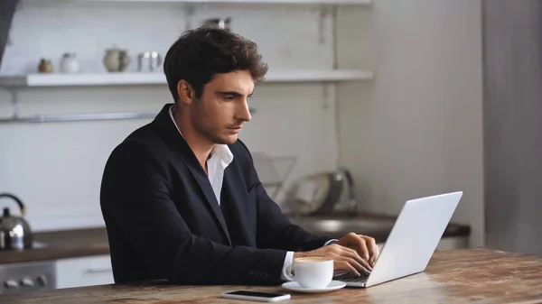 Freelancer using laptop near smartphone and cup of coffee on desk — Fotografia de Stock