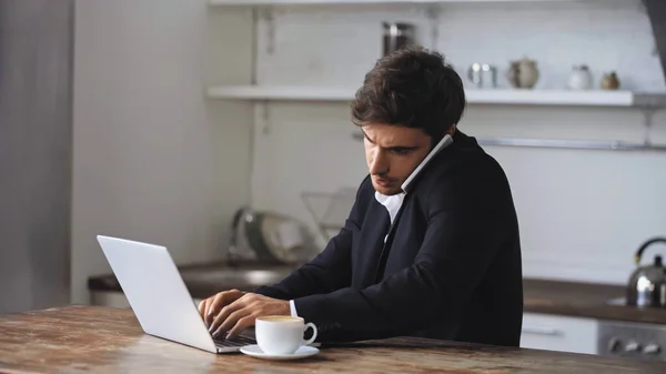 Freelancer talking on smartphone near cup of coffee while using laptop — стоковое фото