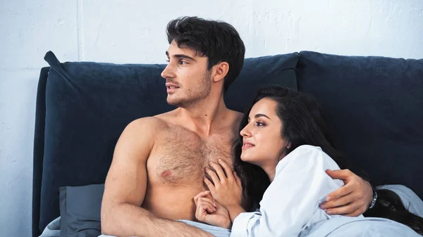 Shirtless boyfriend and smiling girlfriend in white shirt looking away while lying on bed — Fotografia de Stock