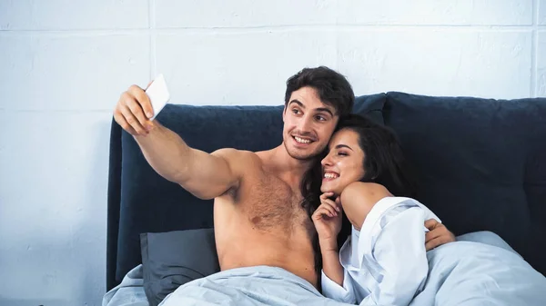 Glücklicher und hemdsloser Mann macht Selfie mit fröhlicher Freundin im Bett — Stockfoto