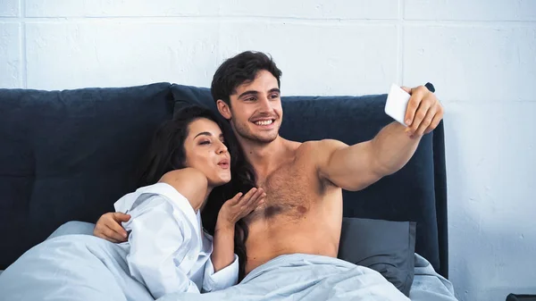 Happy and shirtless man taking selfie with girlfriend sending air kiss while lying in bed — Photo de stock