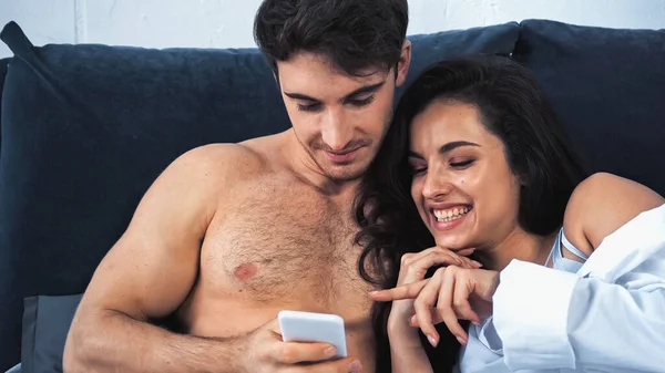 Mulher feliz apontando com o dedo para o smartphone perto de namorado sem camisa no quarto — Fotografia de Stock