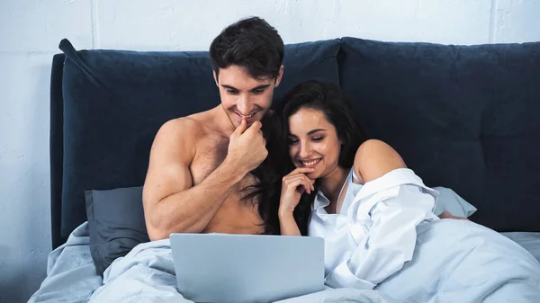 Happy shirtless man and cheerful woman watching comedy movie on laptop in bed — стоковое фото