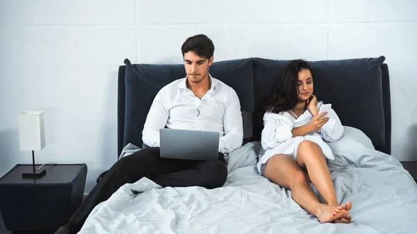 Freelancer using laptop while girlfriend texting on smartphone while resting in bed — стоковое фото