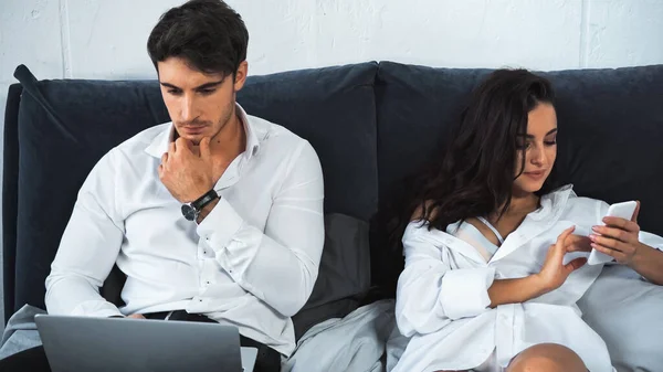 Pensive freelancer using laptop while pretty girlfriend chatting on smartphone while resting in bed — Fotografia de Stock