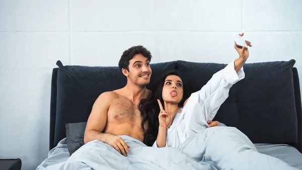 Young woman sticking out tongue while taking selfie with cheerful and shirtless man in bed — стоковое фото