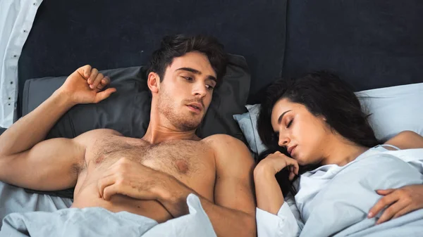 Awakened shirtless man looking at brunette woman with closed eyes sleeping in bed — Foto stock