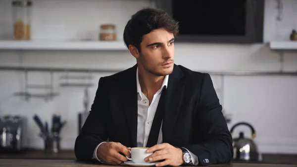 Businessman in formal wear holding cup of coffee while sitting in kitchen — стоковое фото