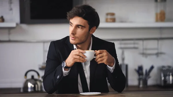 Uomo d'affari in giacca e cravatta in possesso di una tazza di caffè mentre seduto in cucina — Foto stock