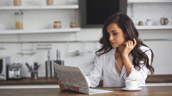 Brünett und hübsche Frau liest Zeitung in der Nähe einer Tasse Kaffee — Stockfoto