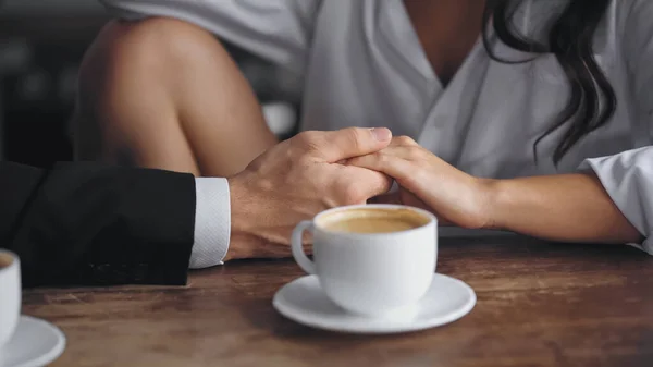 Cropped view of man and woman holding hands near cup of coffee on kitchen table - foto de stock