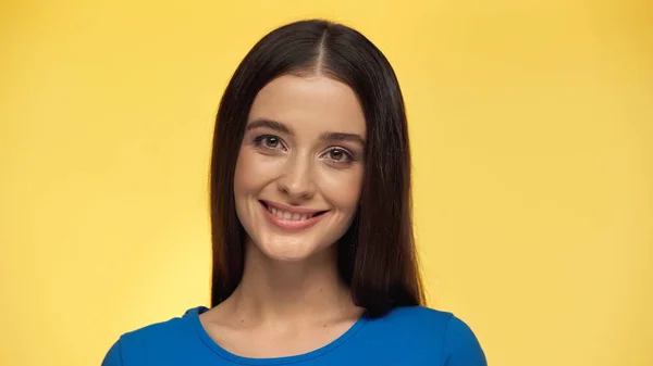 Young brunette woman in blue t-shirt smiling and looking at camera isolated on yellow — Stock Photo