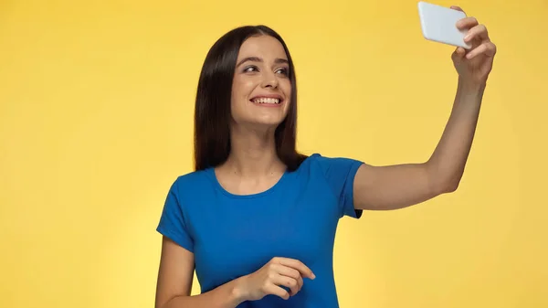 Young brunette woman in blue t-shirt smiling while taking selfie isolated on yellow - foto de stock