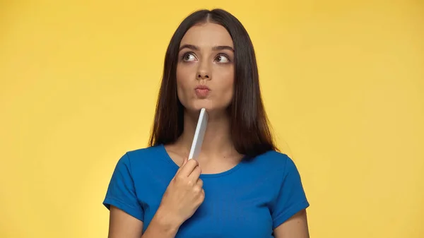 Pensive young woman in blue t-shirt pouting lips and holding smartphone isolated on yellow — Stock Photo