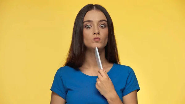 Puzzled young woman in blue t-shirt pouting lips and holding smartphone isolated on yellow — Stock Photo