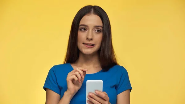 Puzzled young brunette woman in blue t-shirt holding smartphone isolated on yellow - foto de stock