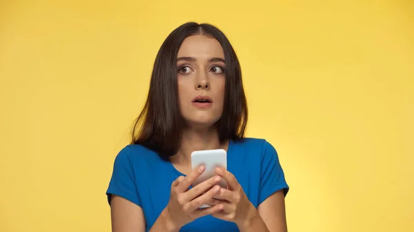 Молода здивована жінка в синій футболці спілкується на смартфоні ізольовано на жовтому — стокове фото