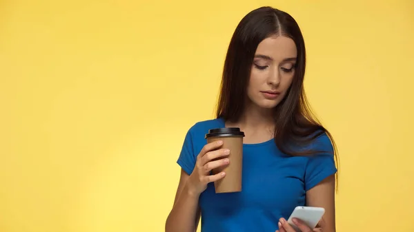 Young brunette woman in blue t-shirt using smartphone and holding coffee to go isolated on yellow — стоковое фото