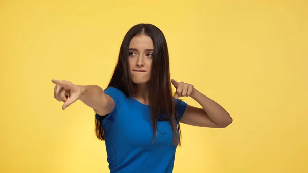 Junge brünette Frau in blauem T-Shirt beißt Lippe und zeigt mit Fingern auf gelb isoliert — Stockfoto