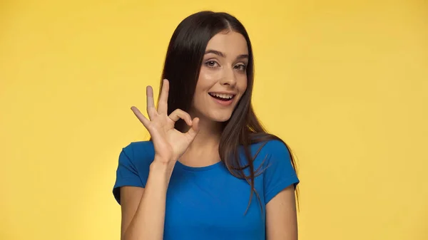 Young and cheerful woman in blue t-shirt showing ok sign isolated on yellow — Fotografia de Stock